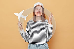 Cheerful woman wearing striped shirt and baseball cap standing isolated over beige background crossing winger praying for good