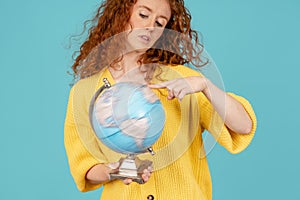 cheerful woman in yellow t-shirt pointing finger at earth globe choosing vacation spot  on blue background