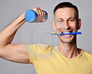 Cheerful young adult man in yellow t-shirt holding toothbrush in his mouth, teeth and a dumbbell in hand in the morning