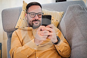 Cheerful young adult mature man happy smiling using smart phone resting lying down on sofa couch at home reading messages on