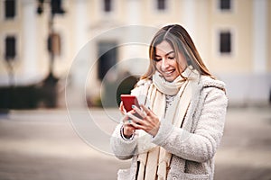 Cheerful young adult woman walking on city street in cold weather and typing a message on her mobile phone - Millennial girl