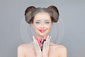 Cheerful young adutl woman with bright pink colour lipgloss holding colorful lipstick in her hands and lookibg at camera on grey