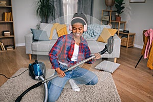 Cheerful young african american female in headphones playing at imaginary guitar as vacuum cleaner in living room