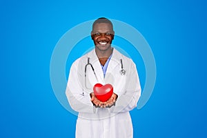 Cheerful young african american man doctor in white coat hold red heart in hands