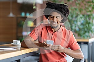 Cheerful Young African American Man Using Smartphone While Relaxing In Cozy Cafeteria