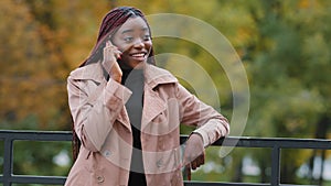 Cheerful young african american woman standing outdoors on autumn day happily chatting on cellular telephone. Smiling