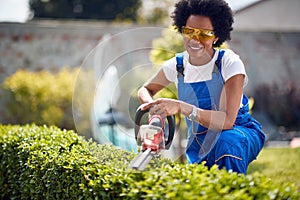 Cheerful young african american woman using electrinc trimmers for cutting hedge outdoors in the yard, working in the garden