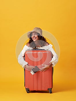 Cheerful young african woman dressed in summer clothes holding passport with money while hug with suitcase isolated over yellow