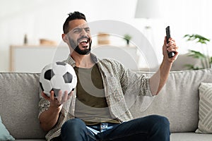 Cheerful young Arab guy watching soccer game on TV, holding ball and remote control, sitting on couch at home