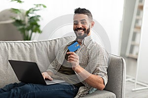 Cheerful young Arab man with laptop and credit card sitting on couch, shopping in online store from home