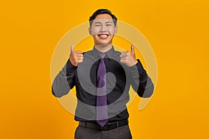 Cheerful young Asian businessman showing thumbs up gesture isolated on yellow background