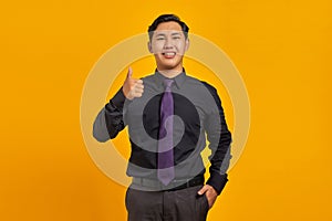 Cheerful young Asian businessman showing thumbs up gesture isolated on yellow background