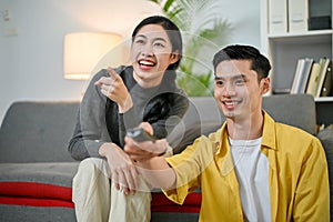 Cheerful young Asian couple are laughing while watching TV together in the living room