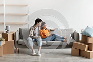 Cheerful young Asian couple using laptop on couch among carton boxes, planning design of their new apartment, copy space