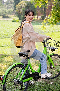 A cheerful young Asian female college student is riding her bike in a park, on her way to campus