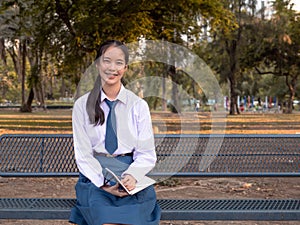 Cheerful young Asian girl student in school uniform Thailand style holds the books in his hands and sitting at the park, Lifestyle