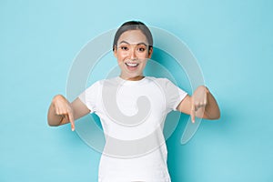 Cheerful young asian lady in white t-shirt pointing fingers down and smiling excited, looking upbeat while demontrating