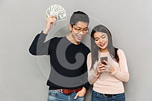 Cheerful young asian loving couple holding money and mobile phone.