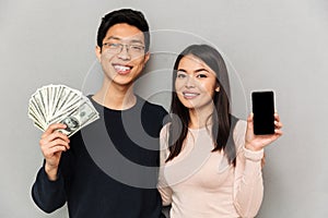 Cheerful young asian loving couple holding money and showing display of mobile phone.