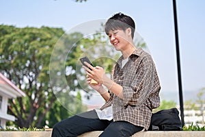 A cheerful young Asian male college student sits on a stone bench in a park, using his smartphone