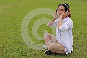 Cheerful young asian man listening to music with headphone in green grass with copy space background.