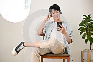 Cheerful young Asian man listening to music through headphones and using his smartphone