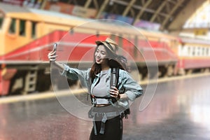 Cheerful young Asian woman traveler with backpack taking a photo or selfie in train station. Travel lifestyle concept