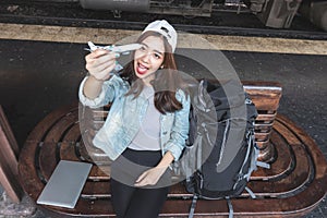 Cheerful young Asian woman traveler with model train sitting on bench waiting for arriving train at station