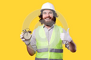 A cheerful young bearded man is showing a thumb up while looking at the camera and holding a little clock near a yellow