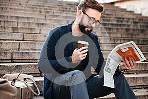 Cheerful young bearded man sitting outdoors on steps reading newspaper