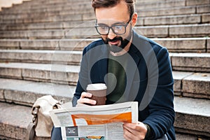 Cheerful young bearded man sitting outdoors on steps reading newspaper