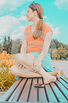 Cheerful young beautiful woman in casual wear sitting on the bench in park and looking away. Girl in orange blouse and cyan skirt