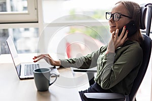 Cheerful young beautiful woman in glasses talking on mobile phone and using laptop with smile while sitting at her working place