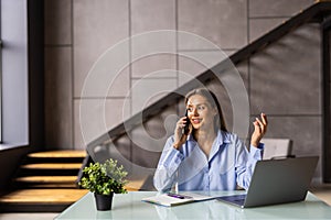 Cheerful young beautiful woman in glasses talking on mobile phone and using laptop with smile while sitting at her working place