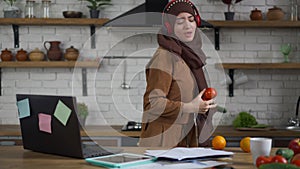 Cheerful young beautiful woman in hijab and headphones singing dancing cooking healthful vegan salad in kitchen