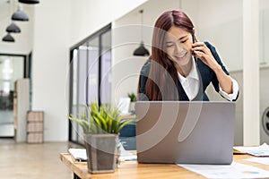 Cheerful young beautiful woman talking on mobile phone and using laptop with smile while sitting at her working place