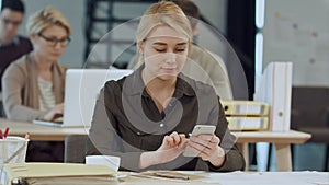 Cheerful young beautiful woman using her smartphone with smile while sitting at her working place