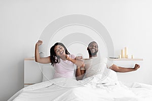 Cheerful young black boyfriend and girlfriend wake up after night sleep and stretch body on bed