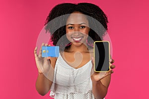 Cheerful young black curly lady shows credit card and phone with empty screen for online shopping
