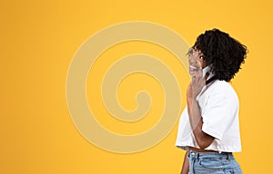 Cheerful young black curly lady in white t-shirt calls by phone, looks at copy space