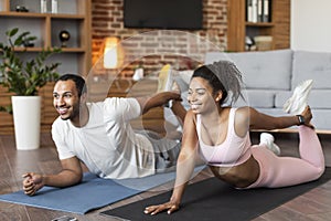 Cheerful young black family in sportswear doing leg stretching exercises on mat on floor, enjoy workout