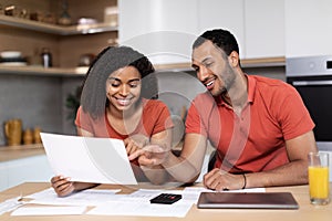 Cheerful young black female in red t-shirt shows documents to man with pc, check accounts use banking