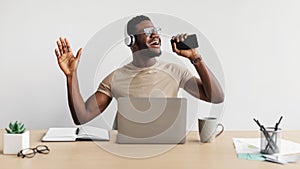 Cheerful young black man listening to music and singing into smartphone while working at desk on white background