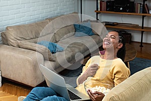 Cheerful young black woman with dreadlocks watching comedy movie on laptop at home, sitting on chair by coffee table, eating