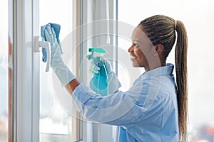 Cheerful young black woman housewife cleaning windows at home