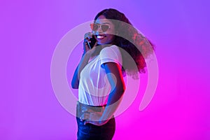 Cheerful young black woman in sunglasses speaking on cellphone, having phone conversation in neon light