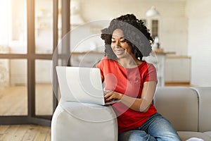Cheerful young black woman using laptop while sitting on sofa at home