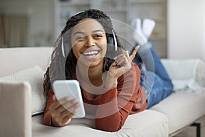 Cheerful Young Black Woman Wearing Wireless Headphones Relaxing With Smartphone At Home