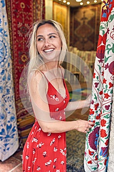 Cheerful young blonde woman in front of carpet and fabric market in wavy red summer dress. Young beautiful woman entering shop.