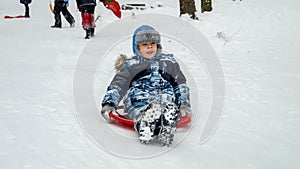 Cheerful young boy sledging down a snowy hill on his plastic sleds, exuding pure joy and enthusiasm. Holiday season and the fun